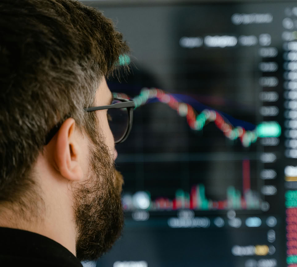 A bearded man with glasses examines trading charts on a computer screen, indicating financial analysis.